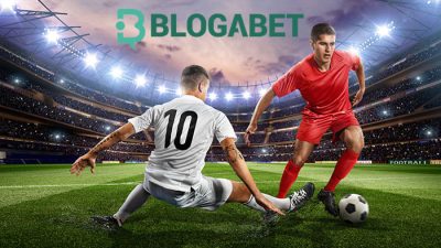 Two soccer players in action on a well-lit stadium field, one in a white jersey sliding to tackle a ball from the other player in a red jersey, with the Blogabet logo prominently displayed above.