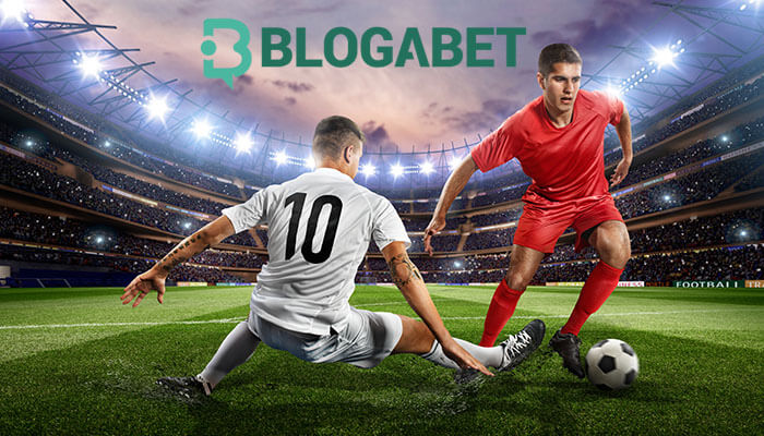 Two soccer players in action on a well-lit stadium field, one in a white jersey sliding to tackle a ball from the other player in a red jersey, with the Blogabet logo prominently displayed above.