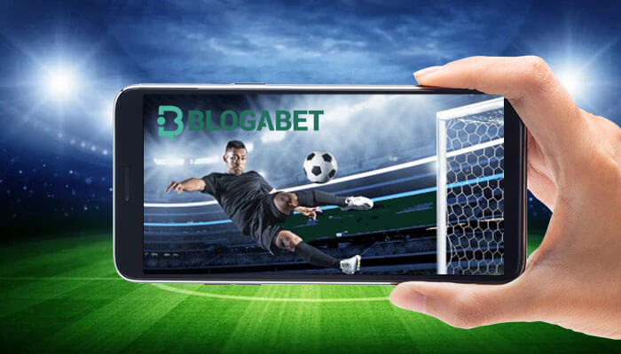 A hand holding a smartphone displaying a soccer player kicking a ball towards a goalpost inside a stadium, with the "BLOGABET" logo visible on the phone screen