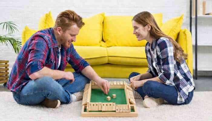 How to set up the Shut the Box game - Live casino Magic - Live CasinoMagic A couple enjoying a friendly match of the Shut the Box game, sitting on the floor with a wooden game board featuring numbered tiles and dice, while a yellow couch is visible in the background.