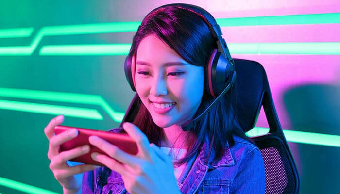The phone has become the default table - Live Casino Magic - Live CasinoMagic Smiling female gamer wearing headphones while playing on smartphone with neon lights, showcasing immersive Digital Gaming experience